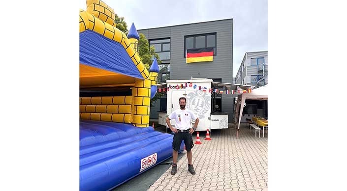 Ein Mann steht vor einer Hüpfburg und einem Gebäude mit deutscher Flagge, dekoriert mit internationalen Flaggen.