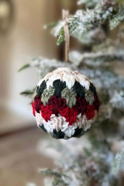 Close-up of a red, green, cream, and white crocheted ornament hanging on a snow-dusted tree branch; no text visible. Free easy crochet Christmas Bauble pattern