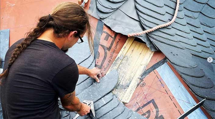 Ein Arbeiter installiert oder repariert eine Schieferdachbedeckung, während er ein Werkzeug hält. Die Hände und einige Details der Dachunterlage sind sichtbar.
