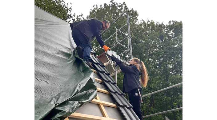 Zwei Arbeiter decken ein Dach mit Dachziegeln; einer reicht dem anderen eine Ziegel, während sie auf einem Gerüst arbeiten.