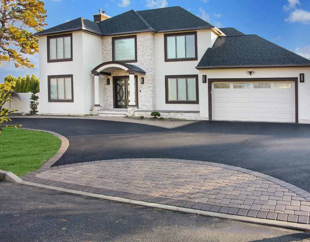 A modern two-story house with white walls, black trim, and a dark roof. The property features a large driveway and a garage, framed by a well-manicured lawn and trees.