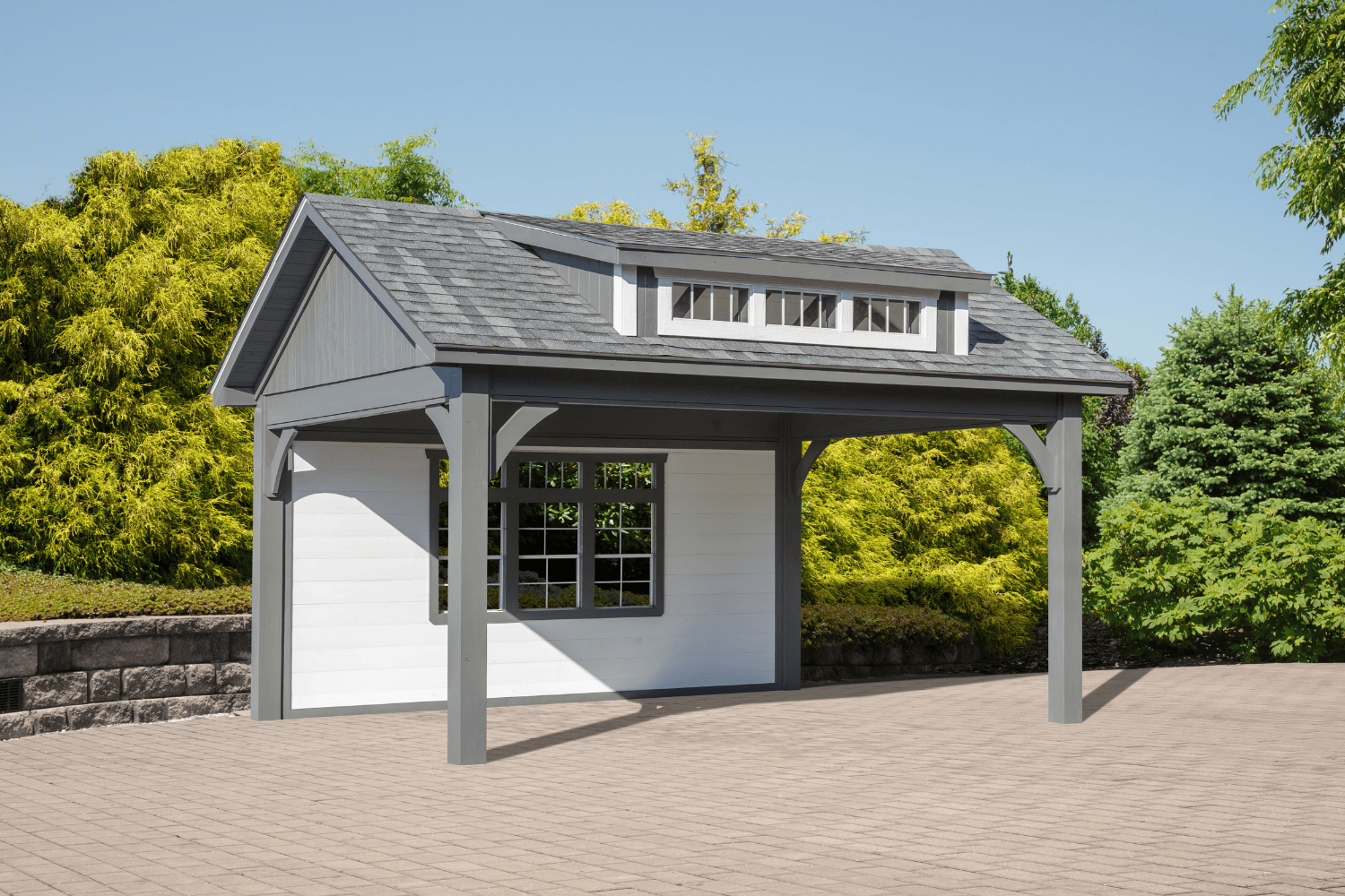 A small, gray and white carport with a gabled roof and dormer stands on a paved area surrounded by trees and shrubs, offering a stylish alternative to traditional pergolas or pavilions.
