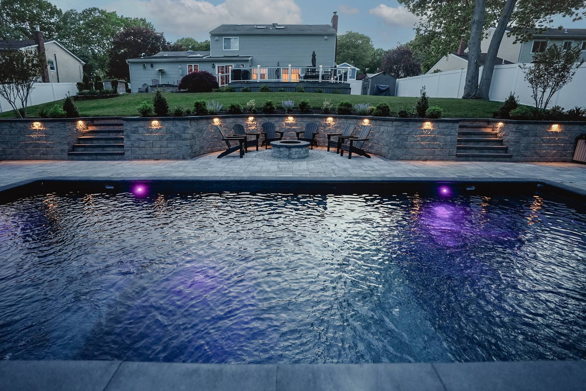 Backyard view featuring a swimming pool with purple lights, stone patio, fire pit with chairs, retaining wall, and a two-story house in the background.