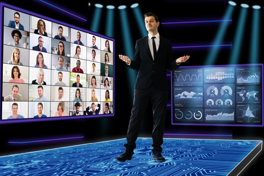 A businessman in a suit stands on a futuristic digital platform with a virtual meeting screen on one side displaying a grid of diverse participants and a data dashboard on the other showcasing charts and graphs. The scene is illuminated with spotlights, symbolizing a high-tech, hybrid environment for remote collaboration, virtual conferences, and data-driven presentations.
