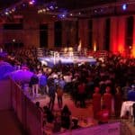 Ein Foto einer Menschenmenge in der Halle, die um einen Boxring stehe. Zwei Männer im Ring kämpfen.
