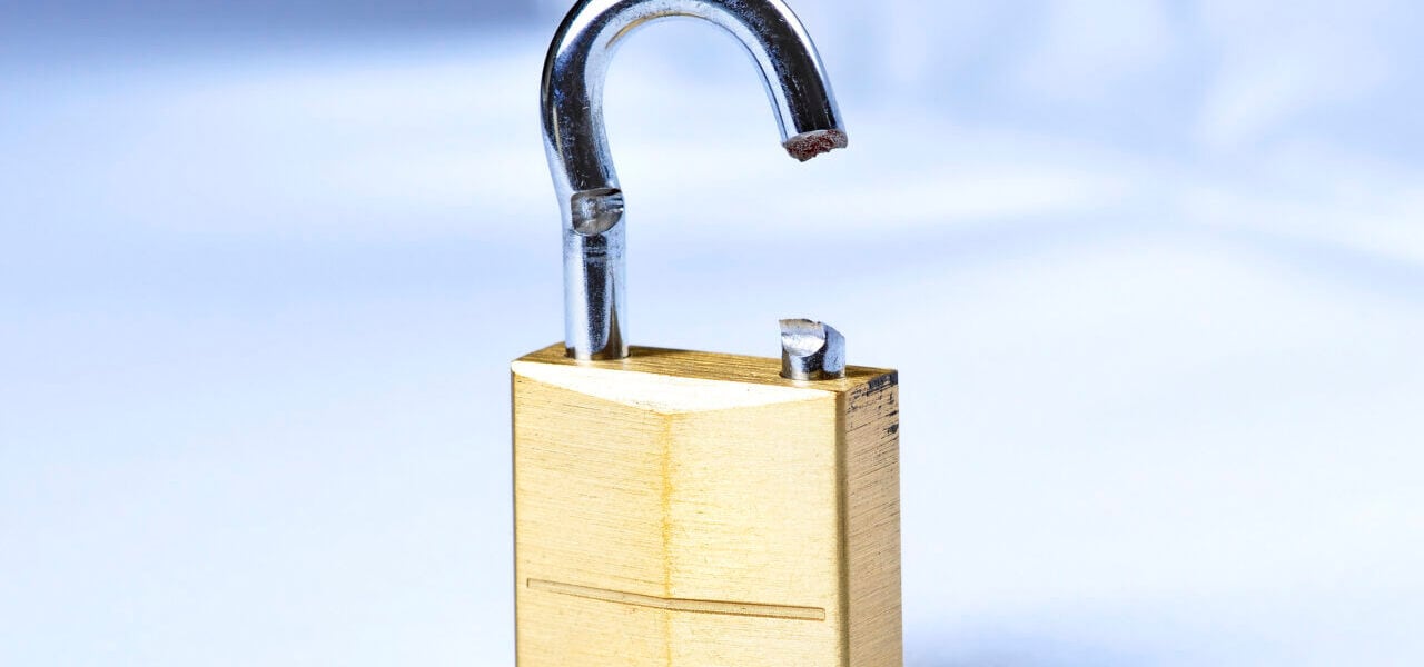 A close-up image of a broken padlock. The padlock, clearly illustrating why a skilled locksmith is essential, is made of brass with a silver shackle, which is bent and snapped on one side, rendering it non-functional. The background is out of focus, making the padlock the focal point of the image.