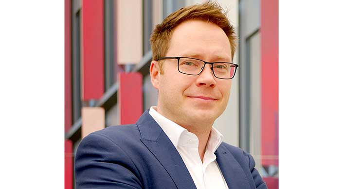 Ein Mann mit Brille und kurzem braunen Haar trägt einen blauen Anzug und ein weißes Hemd, steht draußen vor einem modernen Gebäude.
