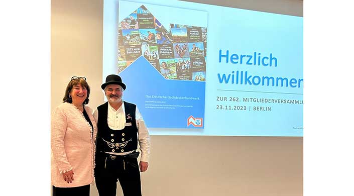 Ein Ausschnitt einer Präsentationsfolie mit dem Titel „Das Deutsche Dachdeckershandwerk“ und Bildern von Dachdeckerarbeiten, teilweise eine Person im weißen Hemd am Rand sichtbar.