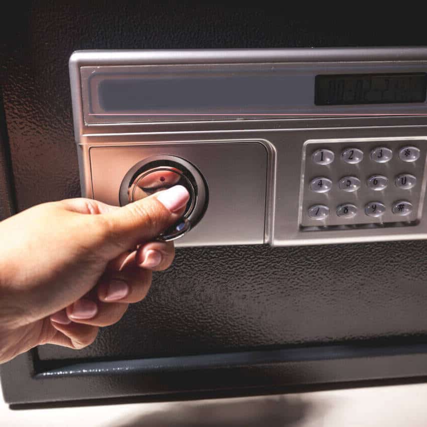 A locksmith is opening a black safe by turning a dial with their left hand. The safe also features an electronic keypad and a small display screen above the dial. Only the hand and part of the forearm are visible in the image.