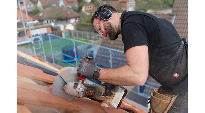 Mann trägt Gehörschutz und Schutzbrille, arbeitet mit einer elektrischen Säge an einem Holzstück im Freien.