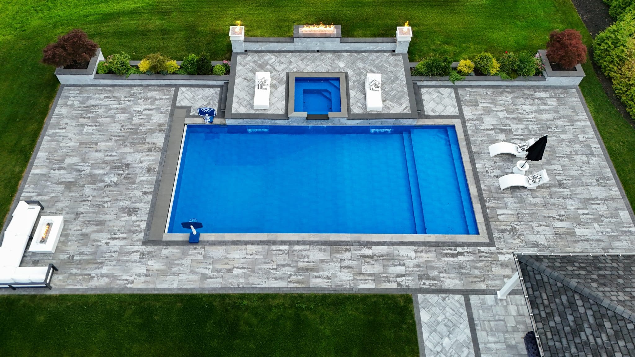 A rectangular outdoor swimming pool with a smaller attached pool, surrounded by gray stone tiles, lounge chairs, and landscaping on a green lawn.
