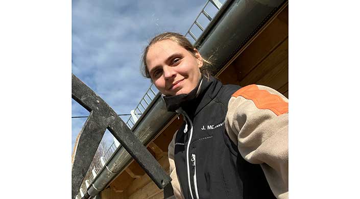 Eine Frau mit braunen Haaren trägt eine schwarze Jacke und steht im Freien vor einem Gebäude mit Metallgeländer.