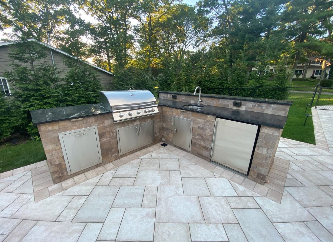 A built-in outdoor kitchen with a grill and sink on a stone patio, surrounded by trees and a house in the background.