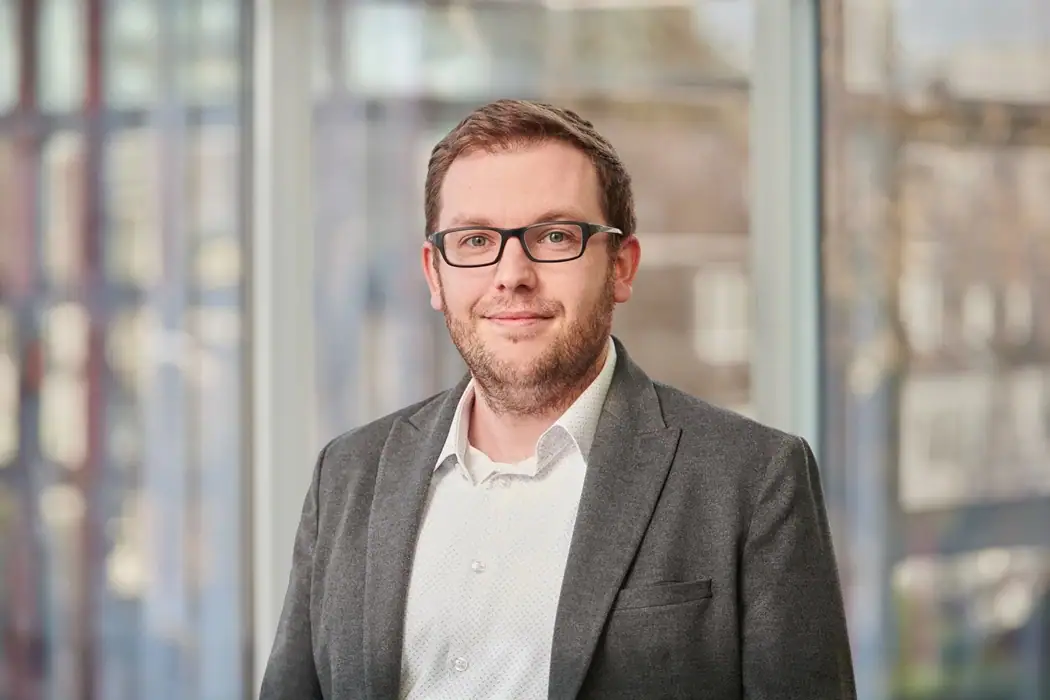 Jan Redecker mit Brille und Bart trägt graues Sakko und weißes Hemd, steht vor einem unscharfen Fensterhintergrund.