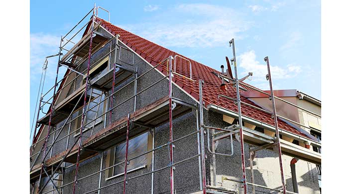 Ein Haus mit Gerüst, grauer Fassade und rotem Ziegeldach, vor blauem Himmel, Renovierungs- oder Bauarbeiten werden durchgeführt.