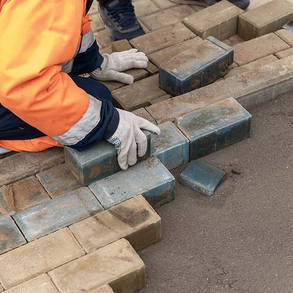 Worker in reflective vest and gloves laying bricks on a pavement.