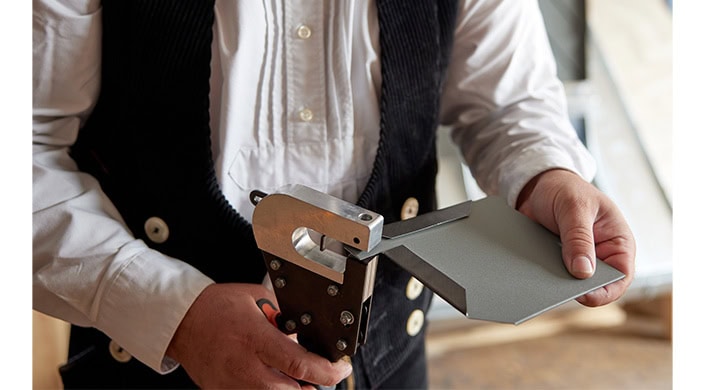 Handwerker hält ein Metallwerkzeug, trägt weiße Hemd und schwarze Weste mit großen Knöpfen.