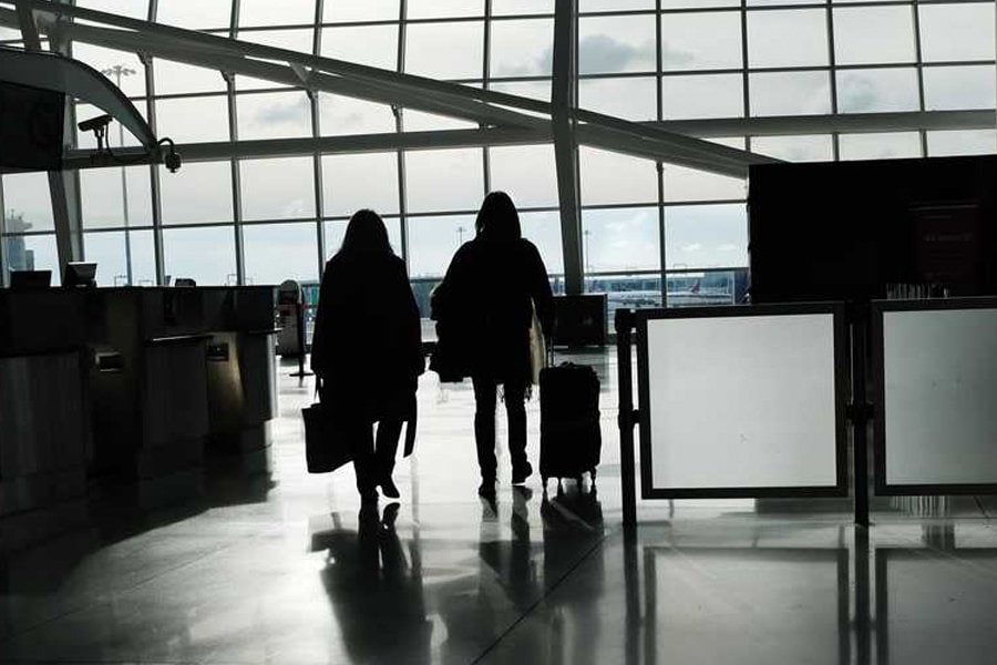 Silhouetted against a large glass window, two travelers walk side-by-side through an airport terminal. The spacious area is illuminated by natural light streaming through the windows, creating reflections on the polished floor. Each traveler carries luggage: one holds a bag, while the other pulls a rolling suitcase. The scene captures the busy yet serene atmosphere of a modern airport.