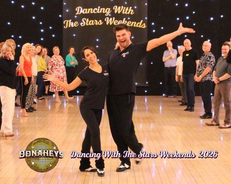 Couple dancing center stage in a studio with a starry backdrop and audience along the walls; sign reads 'Dancing With The Stars Weekends'.