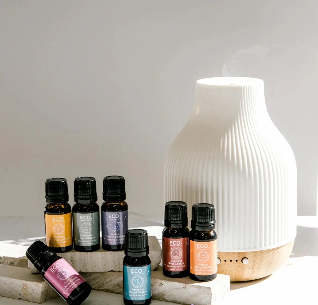 Revive & Thrive Aromatherapy Bundle 4 The Chakra Collection with Diffuser Aromatherapy Bundle features a white ceramic diffuser and seven colorful essential oil bottles, elegantly arranged on stone blocks with steam rising, all set against a neutral background.