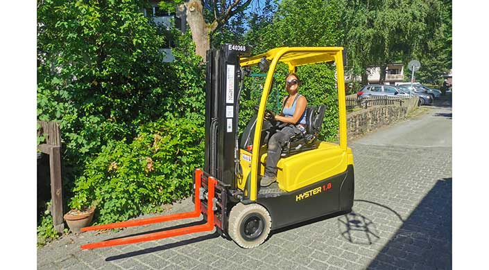 Eine Person fährt bei sonnigem Wetter einen gelben Gabelstapler auf einem gepflasterten Weg, umgeben von grünen Bäumen und Büschen.
