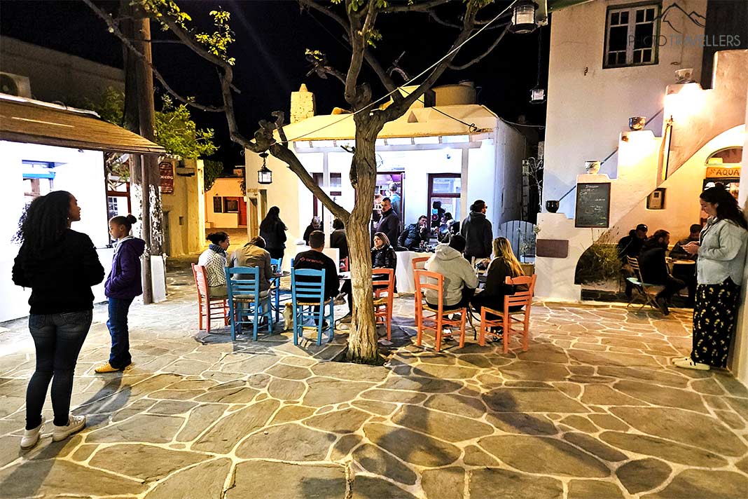 Menschen stehen und sitzen nachts auf einem Platz mit Bäumen und Läden in der Chora von Folegandros