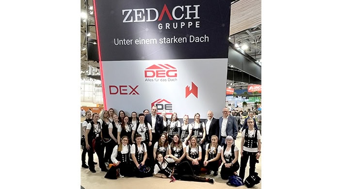Eine Gruppe von Menschen in traditioneller Arbeitskleidung steht vor einem Messestand mit dem Logo der Dach-Gruppe und dem Slogan „Alles für das Dach“.