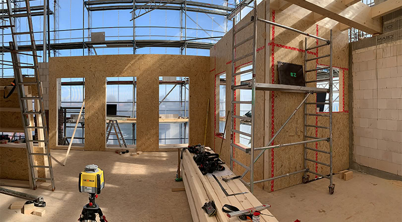 Holzrahmenbau auf einer Baustelle, mit Werkzeugen auf Holzbalken, Gerüst und sichtbarem Fensterrahmen.