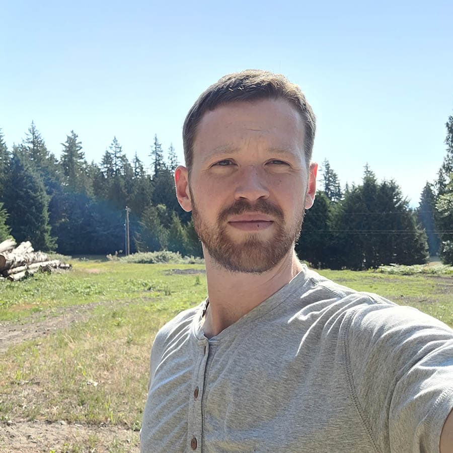 A man with short brown hair and a beard stands outdoors in a grassy area with trees and logs in the background, wearing a light gray shirt.