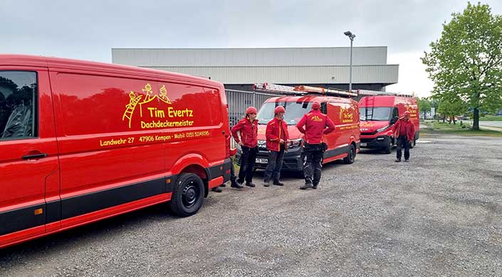 Mehrere Feuerwehrleute in roter Uniform stehen neben roten Einsatzfahrzeugen auf einem Parkplatz vor einem Industriegebäude.