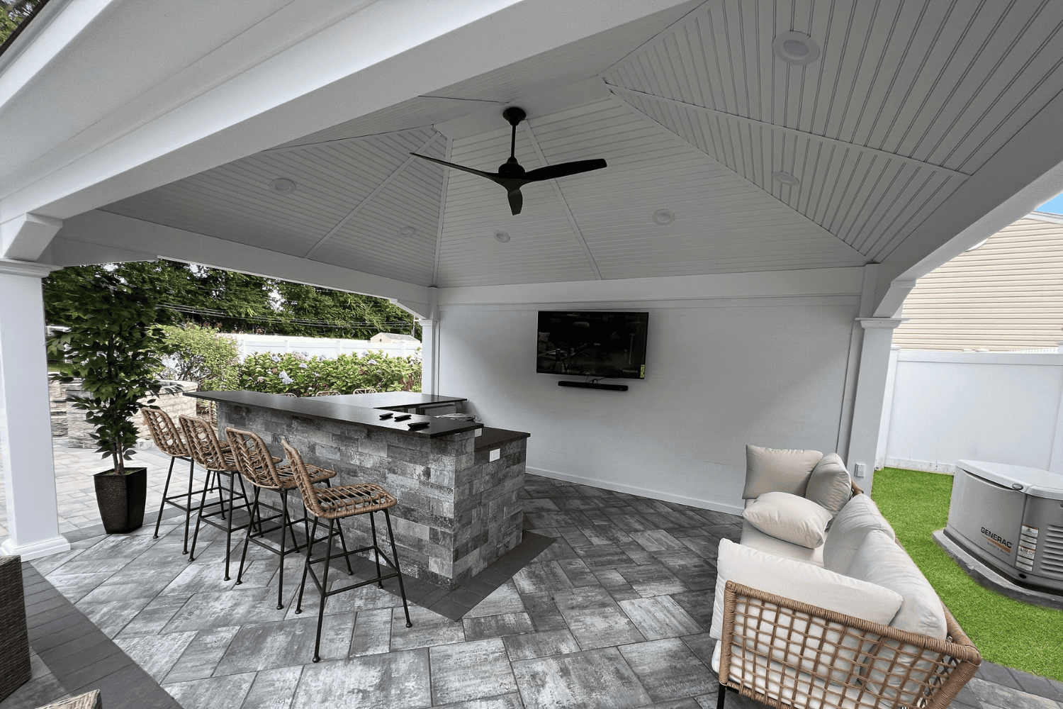 Covered outdoor patio with tiled floor, bar counter with four stools, grill, wall-mounted TV, ceiling fan, cushioned seating area, and a stylish pavilion for added shade and comfort.