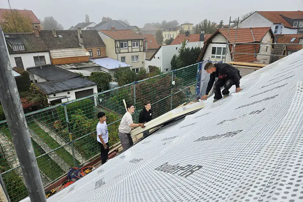Zwei Personen heben Baumaterialien auf einer Baustelle im Freien, im Hintergrund sind Wohnhäuser zu sehen.