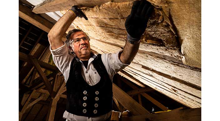 Ein Zimmermann mit traditioneller Kleidung arbeitet mit Holz in einem Dachstuhl aus Holzlatten.
