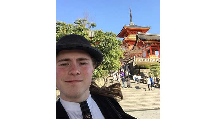 Person mit Hut macht ein Selfie vor einer Treppe, im Hintergrund ein traditioneller japanischer Tempel und Bäume, bei sonnigem Wetter.