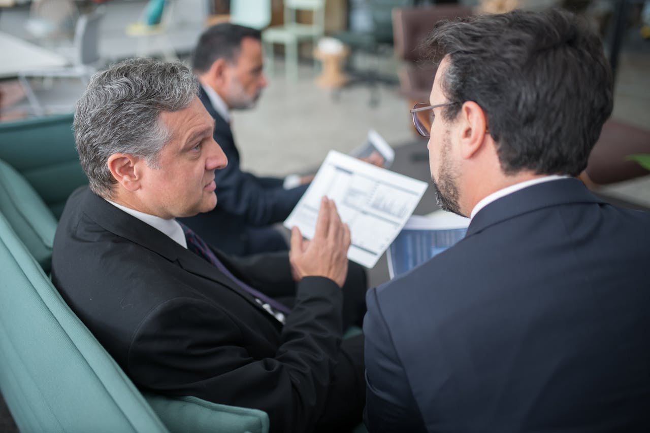men-sitting-on-sofa-having-a-discussion-8353821 Two businessmen engaged in a discussion, examining reports in a modern office environment.