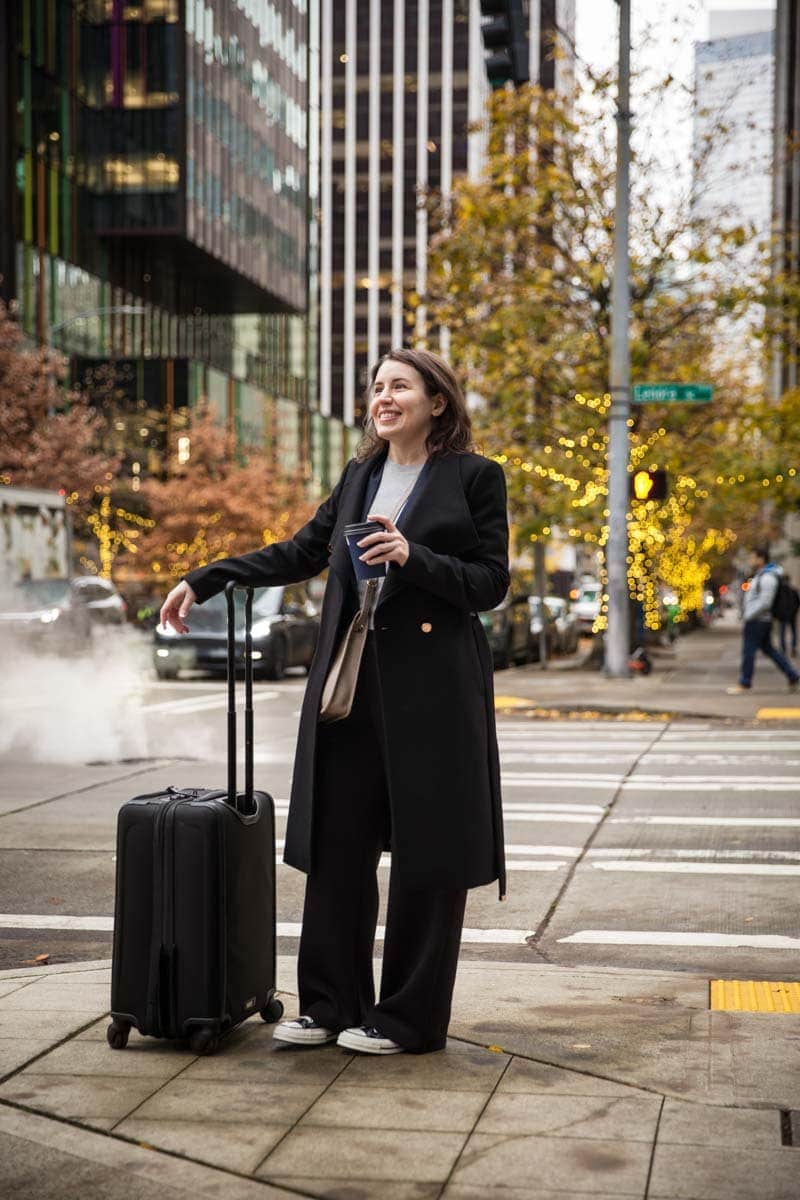 Woman standing on a city street holding a coffee cup, next to a suitcase. Shes wearing a long coat, and buildings and trees with lights are in the background.