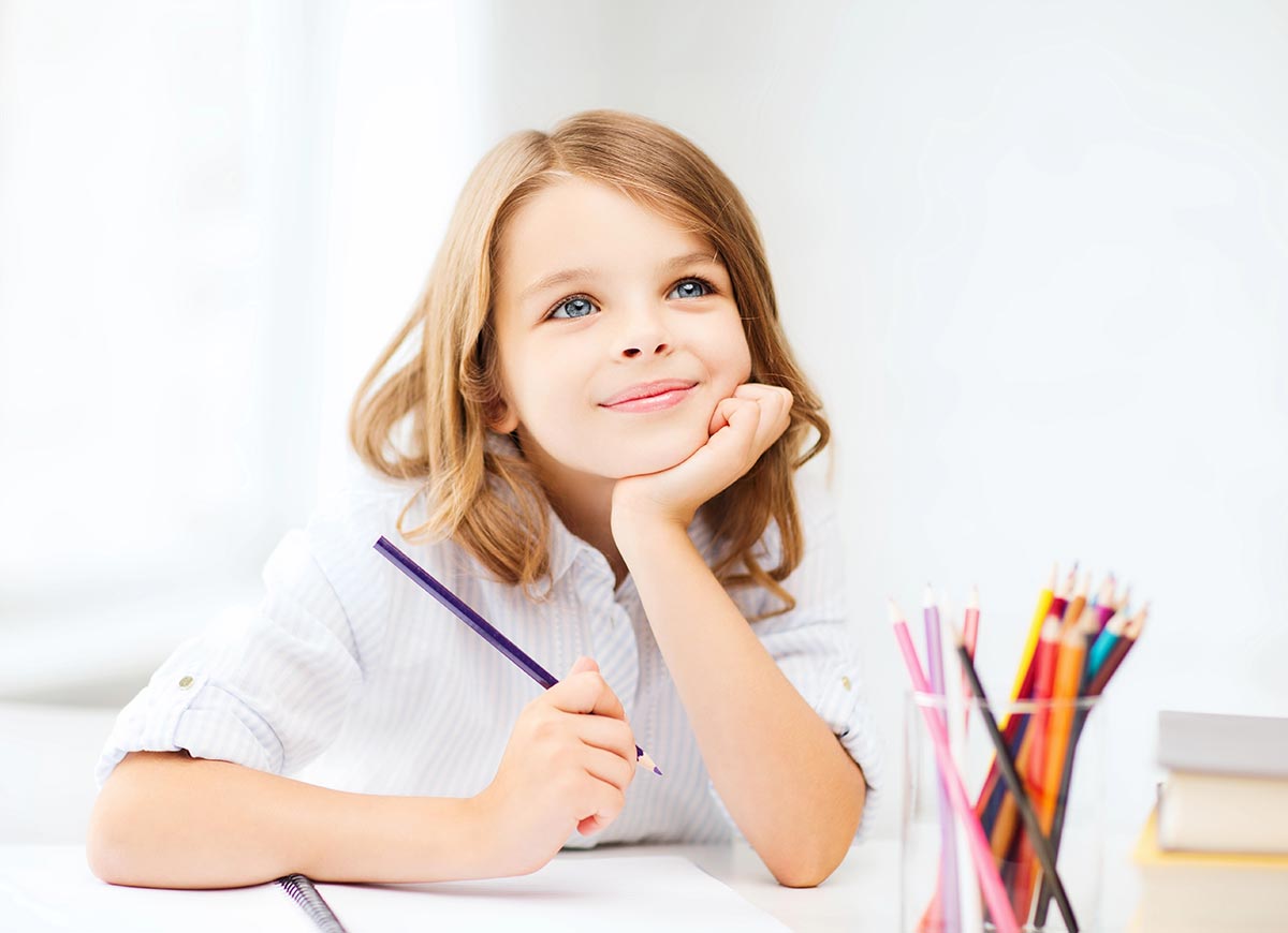 Young child sitting at a desk, holding a pencil and smiling thoughtfully, with colored pencils in a cup nearby.