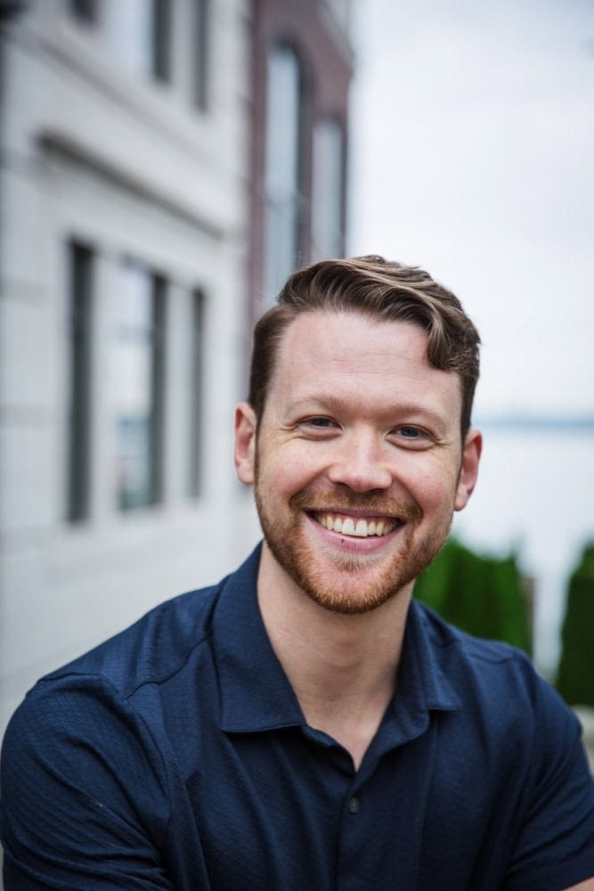 A man with short light brown hair and a beard, wearing a navy blue shirt, smiles while standing outdoors near buildings and greenery.