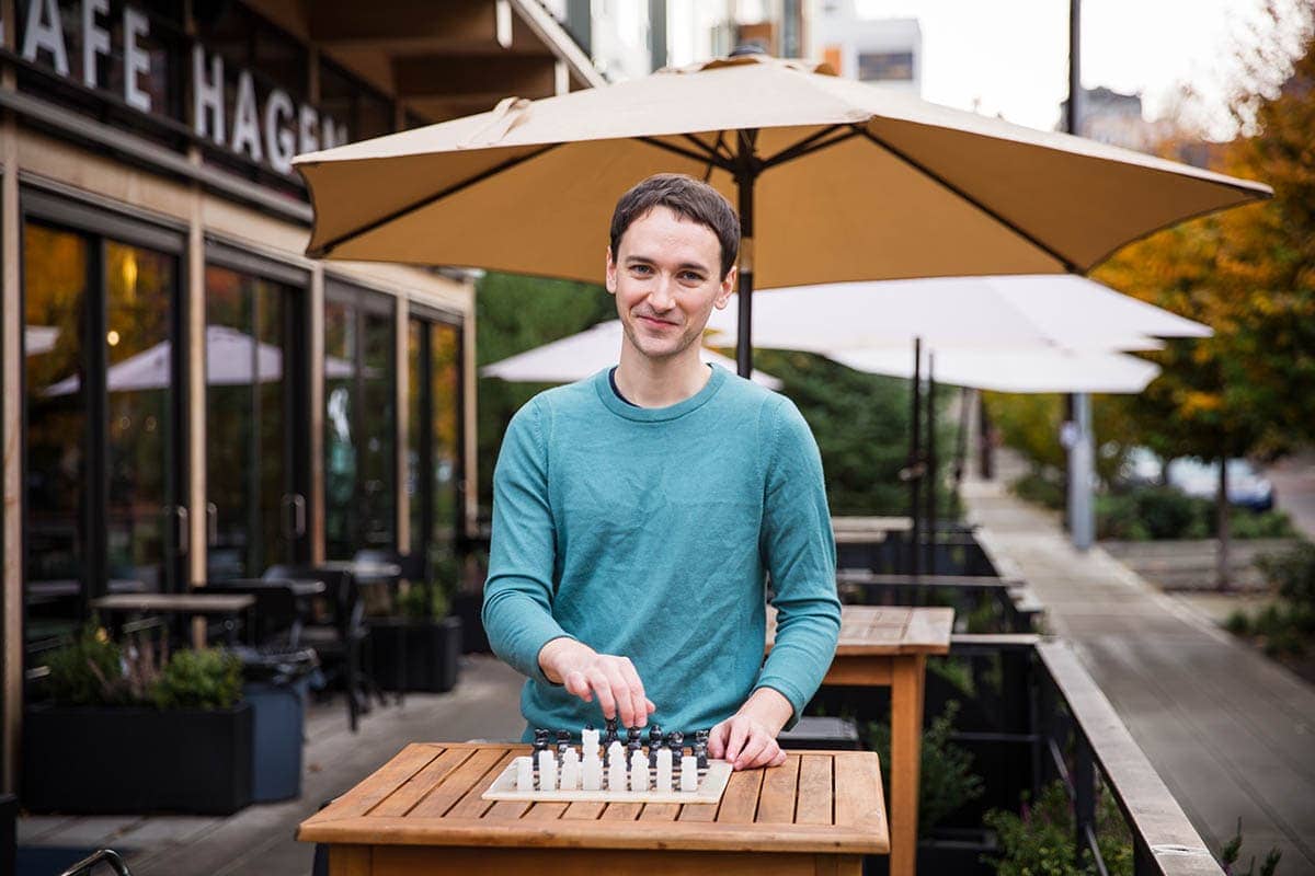 A person in a turquoise shirt is playing chess outside at a wooden table, with large umbrellas in the background.
