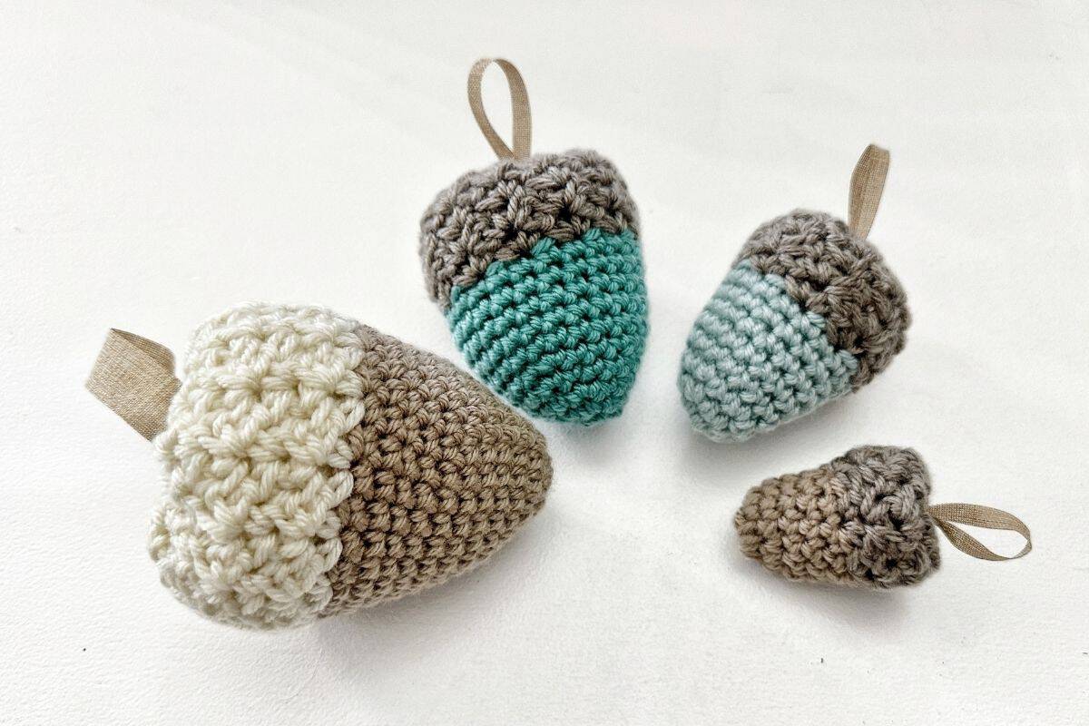 Four crocheted acorn ornaments in beige, brown, and teal with hanging loops are displayed on a white surface. The image promotes a beginner-friendly crochet acorn pattern. Free crochet pattern