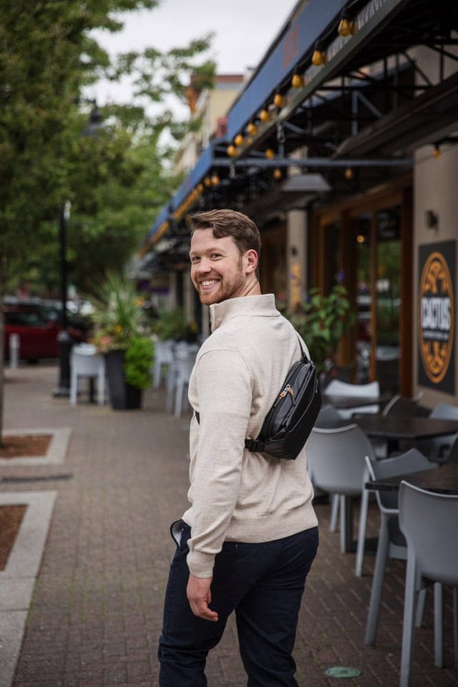 A man with a beard and a beige sweater, wearing a black crossbody bag, looks back and smiles while walking on a sidewalk next to outdoor restaurant seating.
