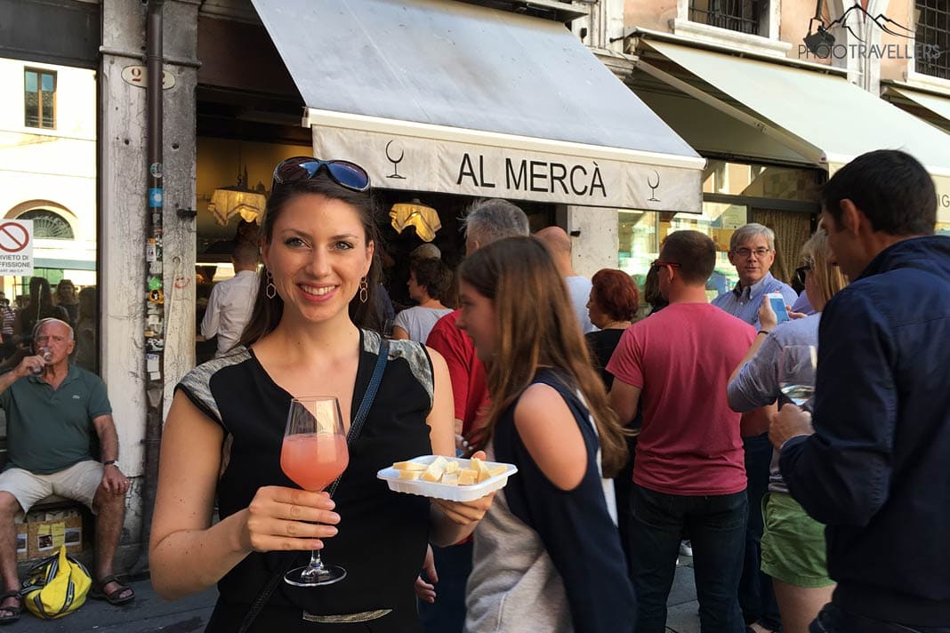 Reisebloggerin Biggi Bauer mit einem Getränk und einem Käse-Snack vor der Bar Al Mercà in Venedig, dahinter viele Menschen