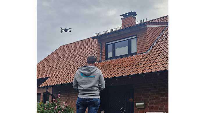 Eine Person steht vor einem Gebäude mit rotem Ziegeldach und großen Fenstern, der Himmel ist bewölkt.