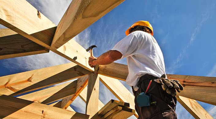Eine Person hämmert einen Nagel in ein Holzbalken, Teil eines Dachstuhls, bei sonnigem Wetter auf einer Baustelle.