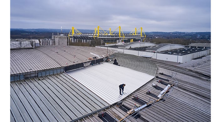 Personen arbeiten auf einem Dach, im Hintergrund ist ein großes Stadion mit gelben Stahlträgern zu sehen.