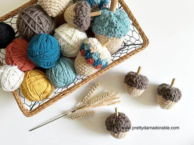 Basket with colorful yarn balls and crocheted acorns on white surface, two acorns and wheat nearby, www.prettydarnadorable.com at bottom. Free crochet pattern