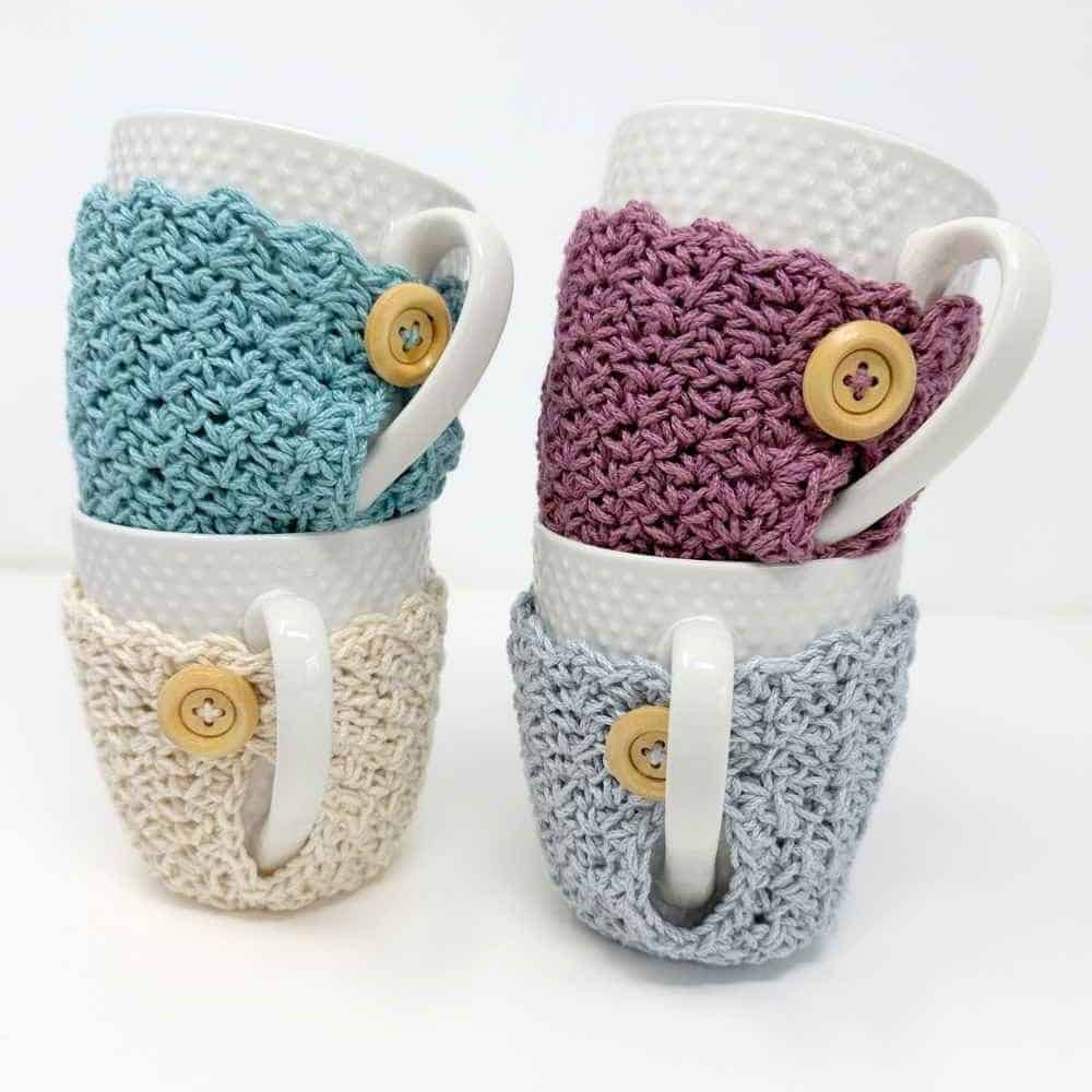 Four white mugs stacked in pairs, each in a beige, blue, or purple crochet cozy with a wooden button on a plain white background. Free crochet mug cozy pattern