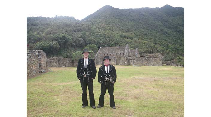 Zwei Männer in traditioneller schwarzer Tracht stehen vor historischen Ruinen und einem bewaldeten Hügel im Hintergrund.