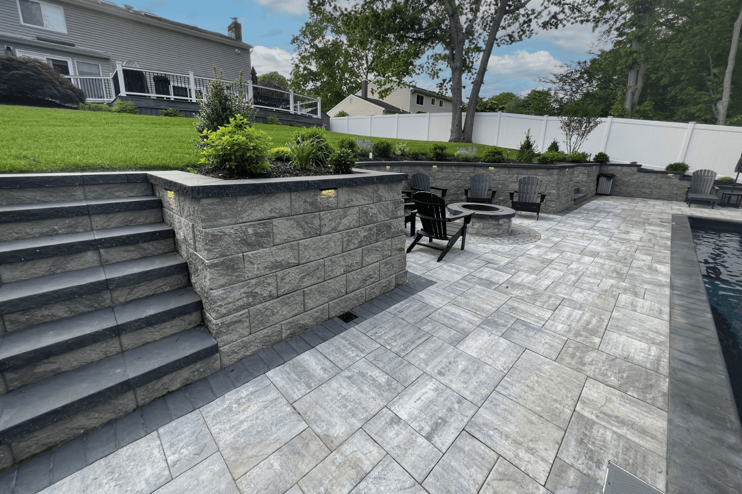 A backyard patio with gray stone pavers, steps leading to an elevated lawn, a fire pit area with chairs, and beautifully landscaped retaining walls under a partly cloudy sky.