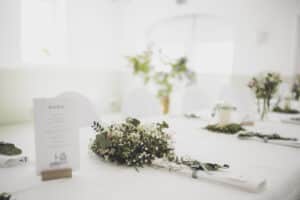 Hochzeitslocation Haus Runde 3 Weiß gedeckter Tisch mit Menü, Blumenschmuck und schlichtem Grün in der Eventlocation Münster.